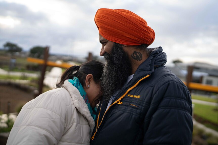 A woman puts her face in her husband's chest. They are standing outdoors with houses in the background. He is wearing a turban.