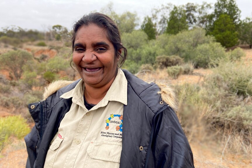 A middle aged indigenous woman wearing navy blue jacket stands in front of native shurbs and bushland smiling at the camera
