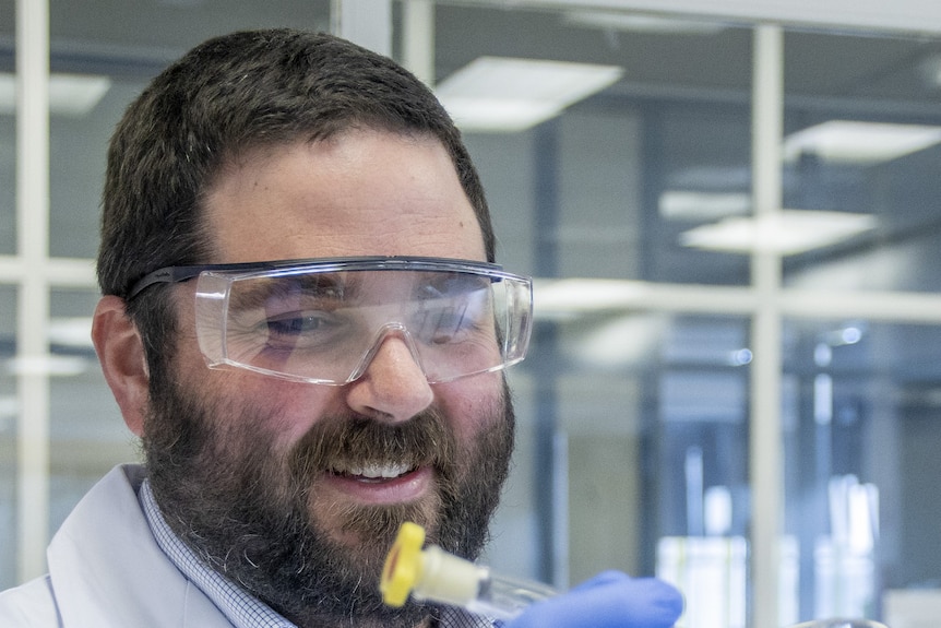Headshot of a brunette man in a lab wearing a white lab coat, blue gloves, protective glasses and a flask of red liquid. 