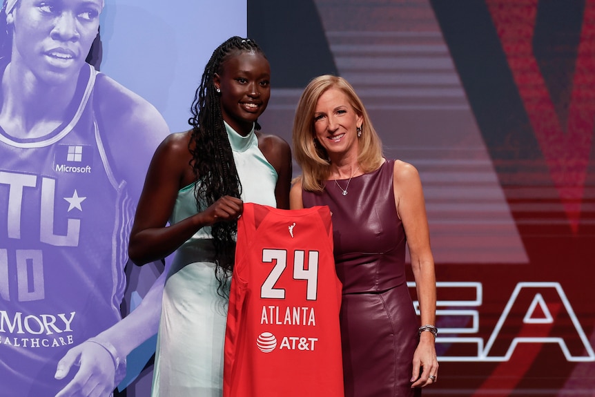 Nyadiew Puoch holds a basketball jersey with "24 Atlanta" on it after being drafted. WNBA commissioner Cathy Engelbert is there.
