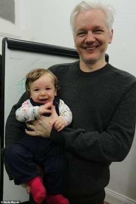 A man with white hair and black clothes holds a smiling baby wearing red socks