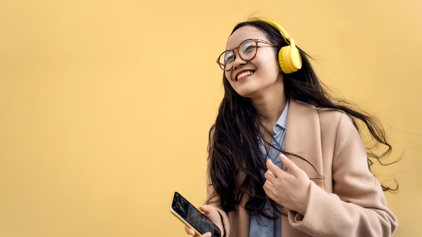 Woman of colour wears yellow headphone, glasses, tan jacket and blue shirt, and holds phone, as she smiles widely and looks up.