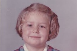 A aged school photo of a smiling primary school aged girl with short blond hair.