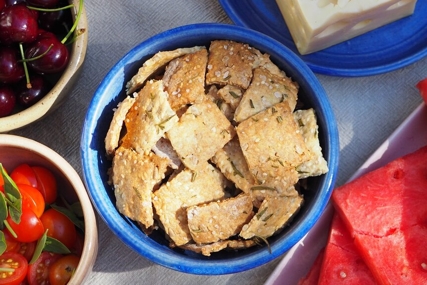 A bowl of sourdough crackers, topped with sea salt, herbs and seeds. Served alongside cheese, fruit, veggies.