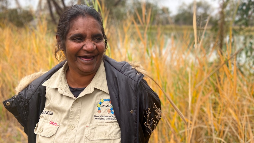 An indigenous woman stands in front of the Murray River smiling with tall yellow reeds separating her from the water