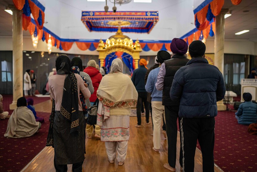 A number of people stand with their backs to the camera in a colourfully adorned Sikh place of worship.