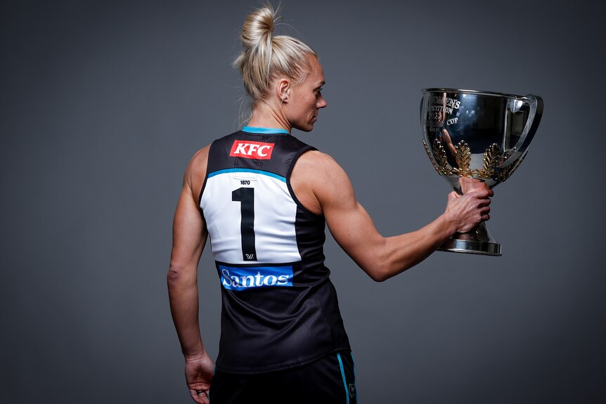 Erin Phillips, with her back to the camera, holds the Premiership Cup to her right and stares into it.