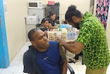 A man sitting down gets an injection from another man wearing a bright green shirt. 