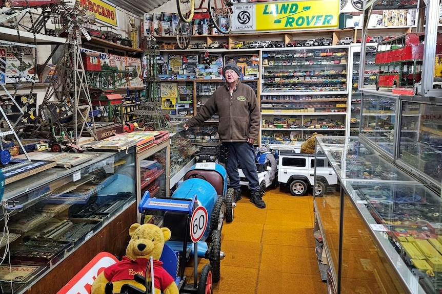 A man in a brown sweater, a black beanie and denim jeans stands in front of children's car toys. 