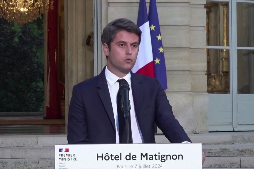 A man at a suit and tie speaking at a microphone with the French and EU flags behind him.