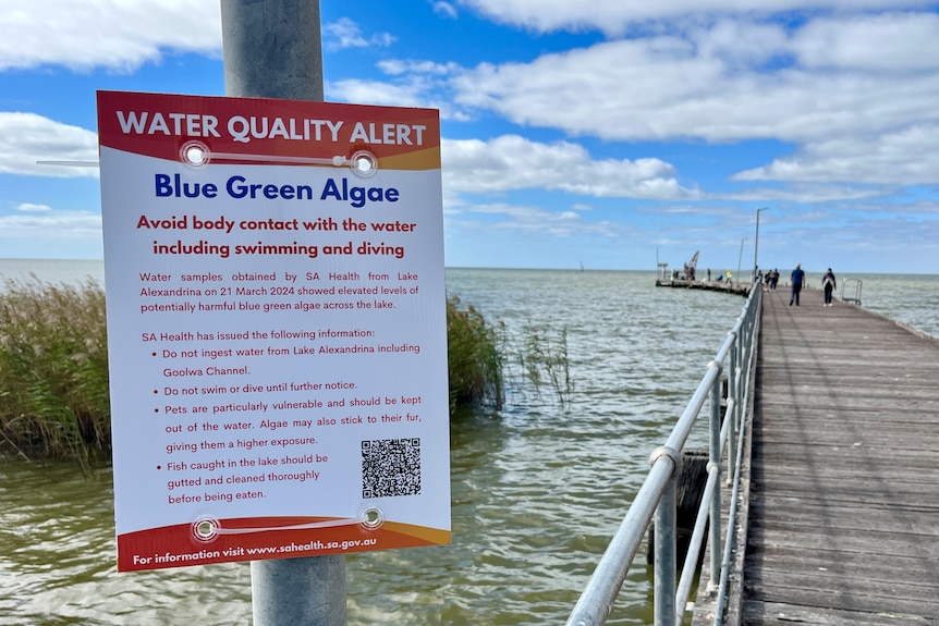 A sign on a pole in front of a boat ramp and jetty warning people to stay out of the water