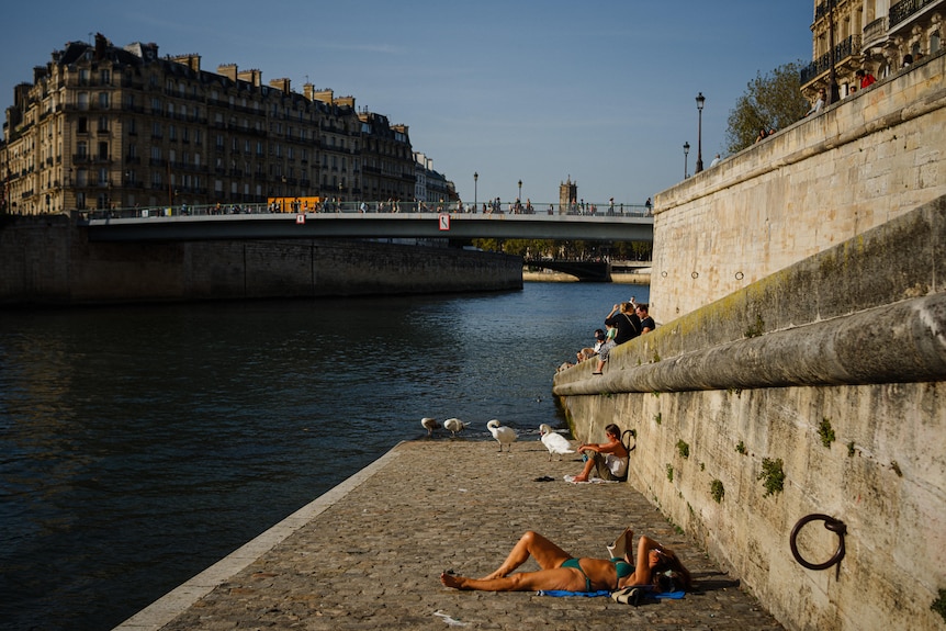 A woman in a green bikini lies on the concrete bank of the Seine river reading, with a view of Paris visible in the background