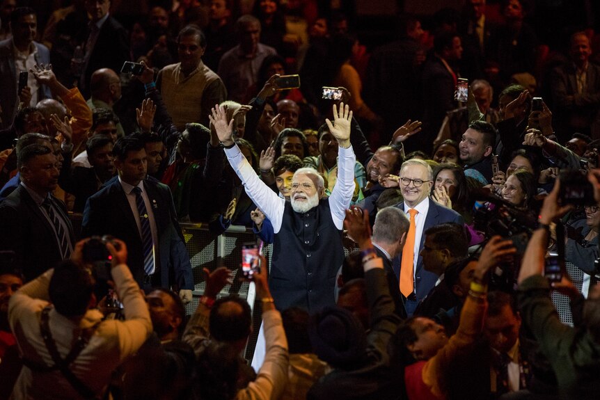 Narendra Modi, standing beside Anthony Albanese at the centre of a crowd, holds up his arms.