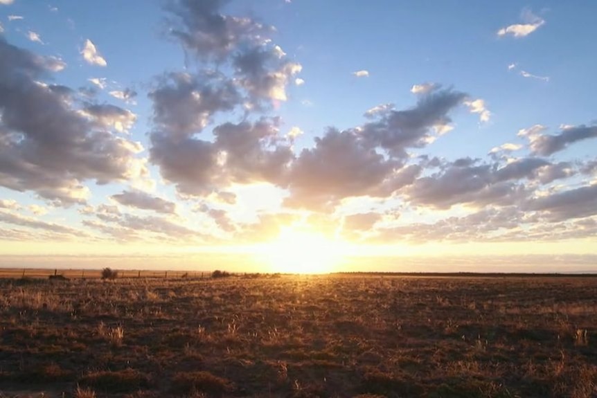 The sun rising over a dry paddock, with blue sky and clouds above.