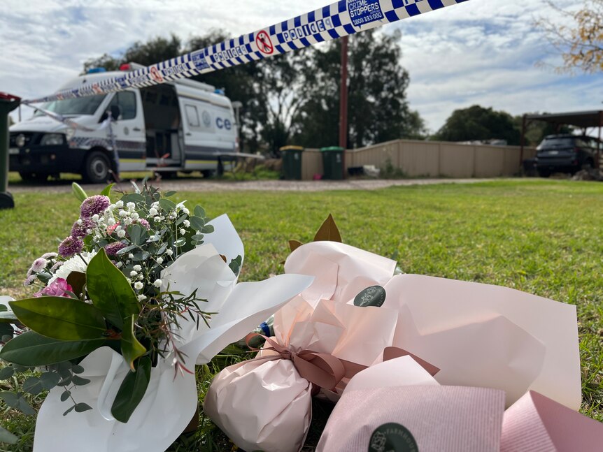 Flowers left outside a house with police tape above the flowers and a van in the background.