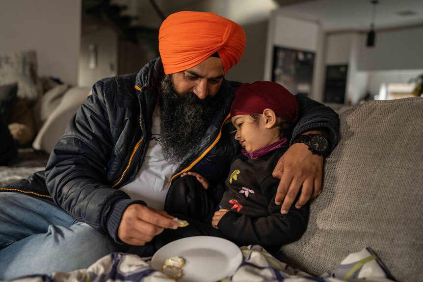 A man wearing a turban puts his arm around a young child sitting next to him on a living room couch.