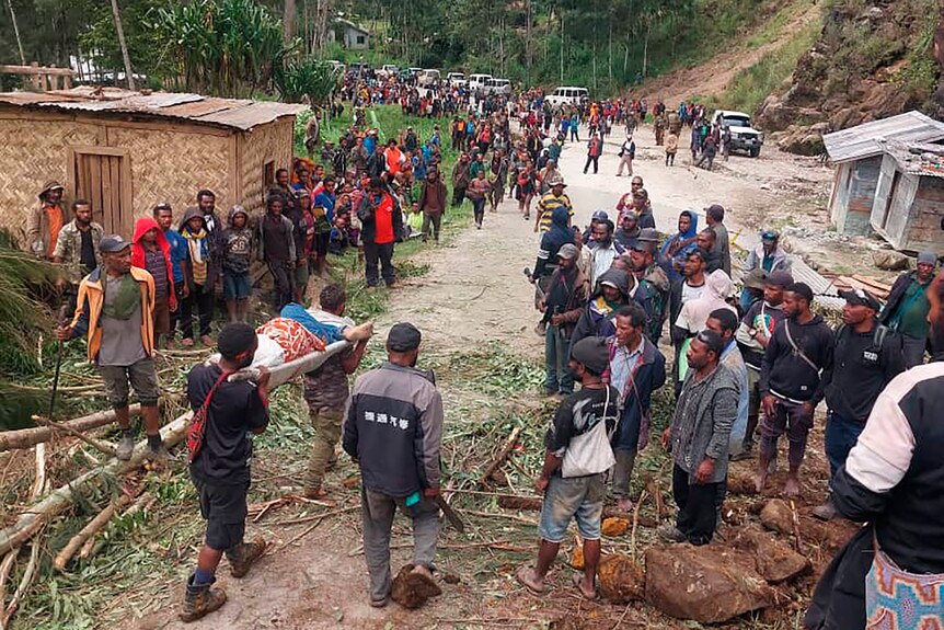  a crowd of people walking on a dirty road, two people carry a stretcher made out of tree branches. 