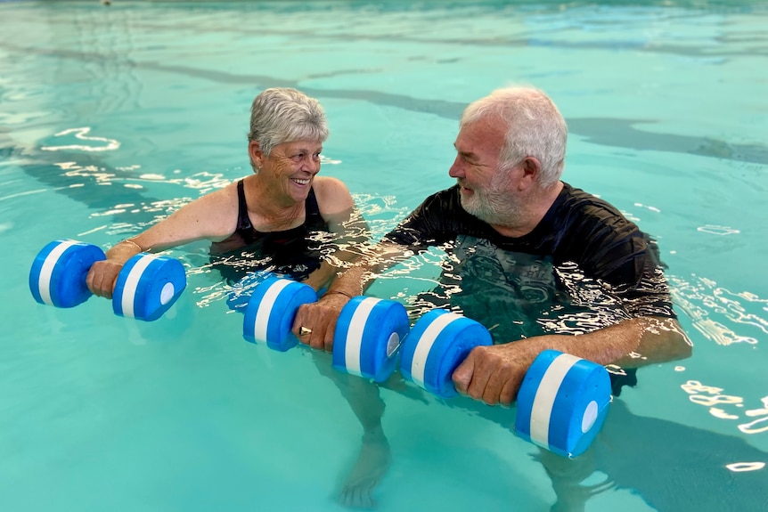 An elderly couple floating in a pool holding blue and white foam dumbbells' smile lovingly at each other.