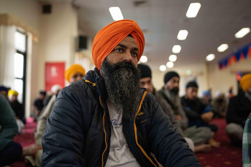 A man wearing a turban sits on the floor looking up at something in front of him, behind him others are doing the same.