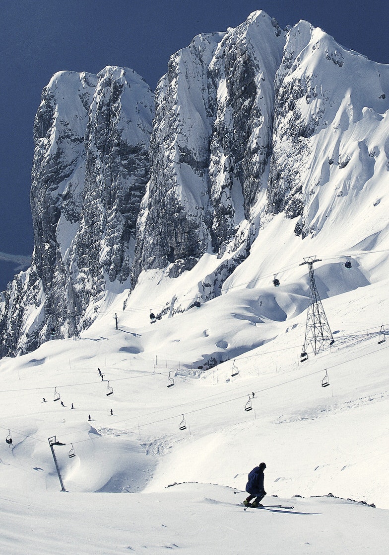 Dramatic rocky outcrops tower above the ski runs. One skier is silhouetted against the snow; many other skiers appear as dots further down the slopes