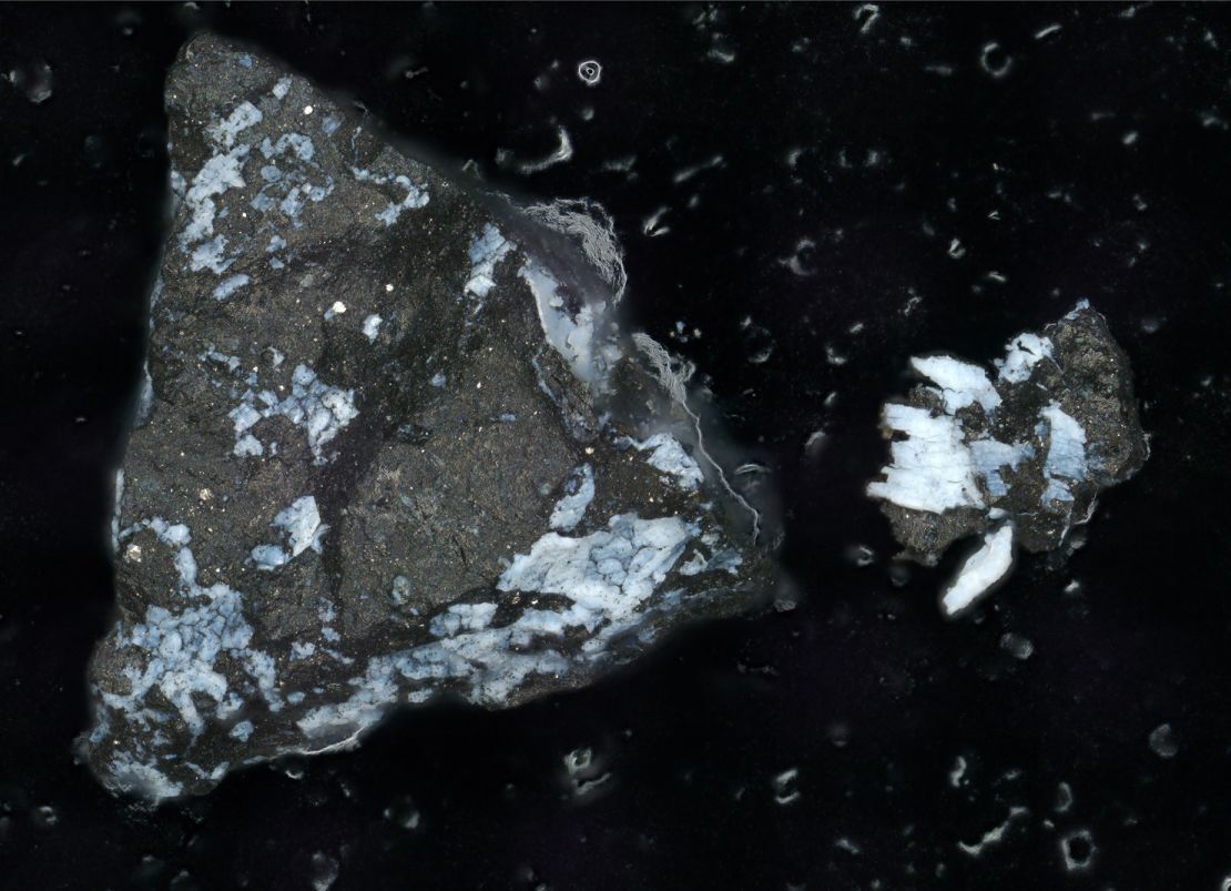 A microscope image shows a dark particle from Bennu, about a millimeter long, with a crust of bright phosphate.