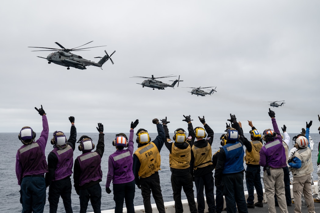 Sailors aboard USS Somerset (LPD 25) wave to CH-53E Super Stallion helicopters from VMM-165 as they fly by the flight deck while underway in the Pacific Ocean.