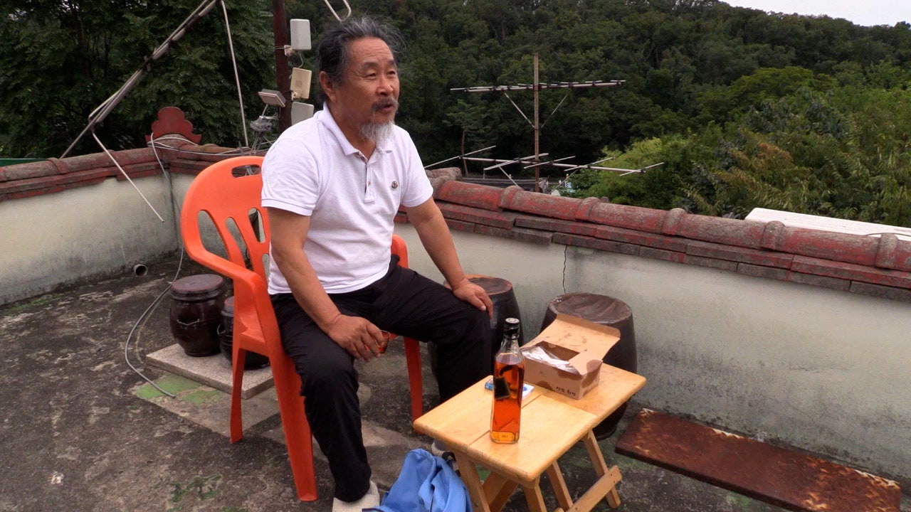 An older man sits in a plastic chair in front of a small table on a balcony.