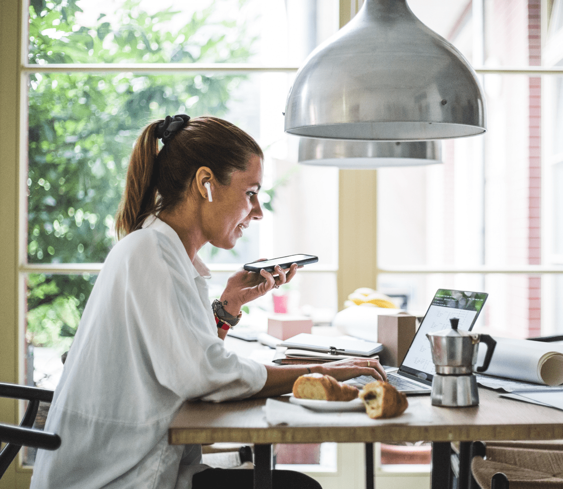 Woman at kitchen table on phone and working on laptop