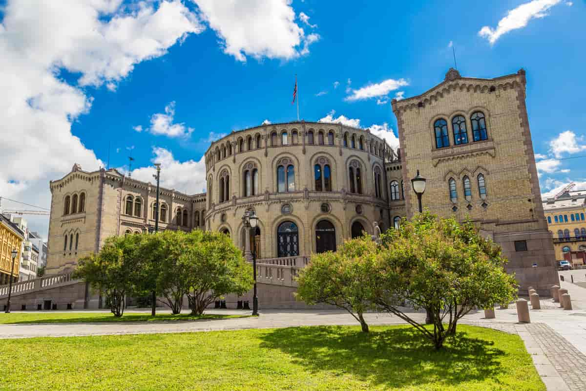 Stortingsbygningen i Oslo. En gammel bygning med buede vinduer og en rund form midt på. Foran bygningen er en plass med gangveier og gressplener med busker. Foto