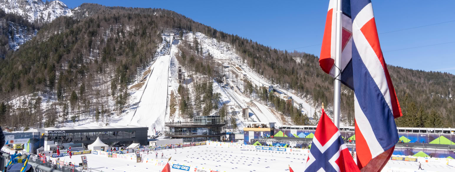 Foto av en skistadion i strålende sol. Flere norske flagg er festet til et gjerde foran startområdet for langrenn. Tre hoppbakker i forskjellige størrelser ligger ved siden av hverandre på en fjellside med grantrær.