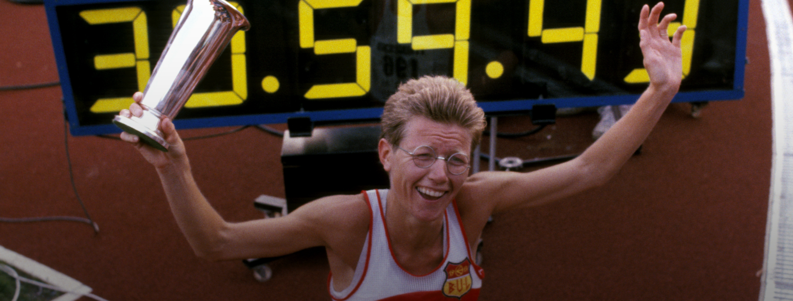 Foto av Ingrid Kristiansen med en pokal i den hånden, sittende på huk foran en stor digital klokke som viser vinnertiden i løpet hennes (30.59.43)