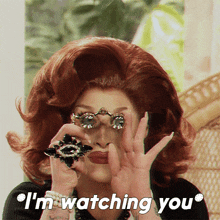 a woman with red hair is holding a pair of binoculars in front of her face and says " i 'm watching you "