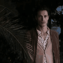 a man in a polka dot shirt and a brown jacket is standing in front of a palm tree