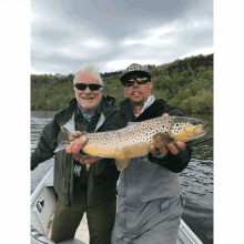 two men in a boat holding a brown trout with one wearing a hat that says ' fishing ' on it