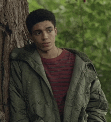 a young man leaning against a tree wearing a green jacket and striped shirt