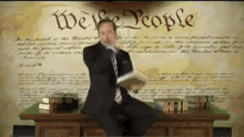 a man in a suit and tie is sitting at a desk in front of a wall with the words we the people written on it