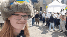 a woman wearing glasses and a fur hat with the word sweden on it stands in front of a crowd of people