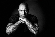 a black and white photo of a man with tattoos on his arms sitting at a table .