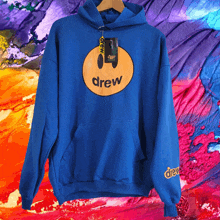 a blue drew hoodie is hanging on a hanger in front of a colorful background