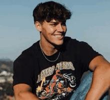 a young man wearing a black t-shirt and a necklace is sitting on a hill and smiling .