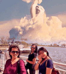 three women are standing in front of a body of water and a statue of a woman with clouds behind them