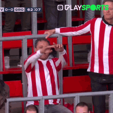 a man in a red and white striped shirt is sitting in a stadium watching a soccer game and the time is 6:07