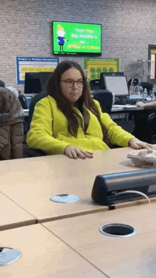 a woman in a yellow jacket sits at a desk in front of a green screen that says " time out "