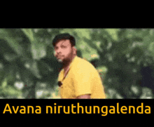 a man in a yellow shirt is standing in front of trees with the words avana niruthangalenda written on the bottom .