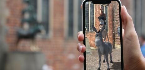 Im Vordergrund sind die Bremer Stadtmusikanten in 3D durch ein Smartphone zu sehen. Im Hintergrund ist die Statue der Stadtmusikanten zu sehen. 
