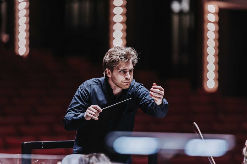 A conductor in a dark shirt holds a baton, intensely directing a performance in a dimly lit venue.