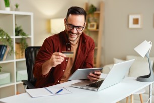 A smiles at his credit card while sitting in front of a laptop and various papers.