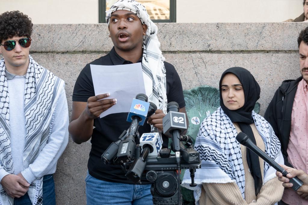 A group of people against a wall and in front of microphones. They are wearing keffiyehs.