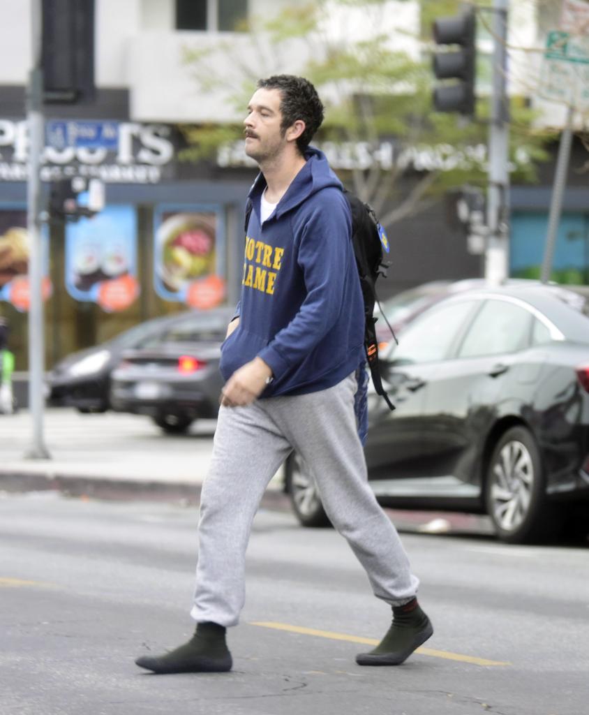 Matty Healy, frontman of The 1975, walking on the street in Los Angeles, dressed in a torn Notre Dame hoodie and grey joggers, heading towards a coffee shop.