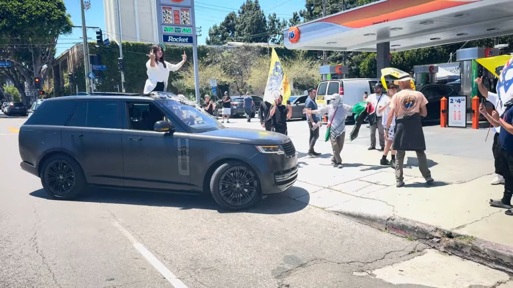 The woman in the vehicle stands up through the sunroof, from where she gives the crowd the middle finger, and makes a shooting gesture with her hand.